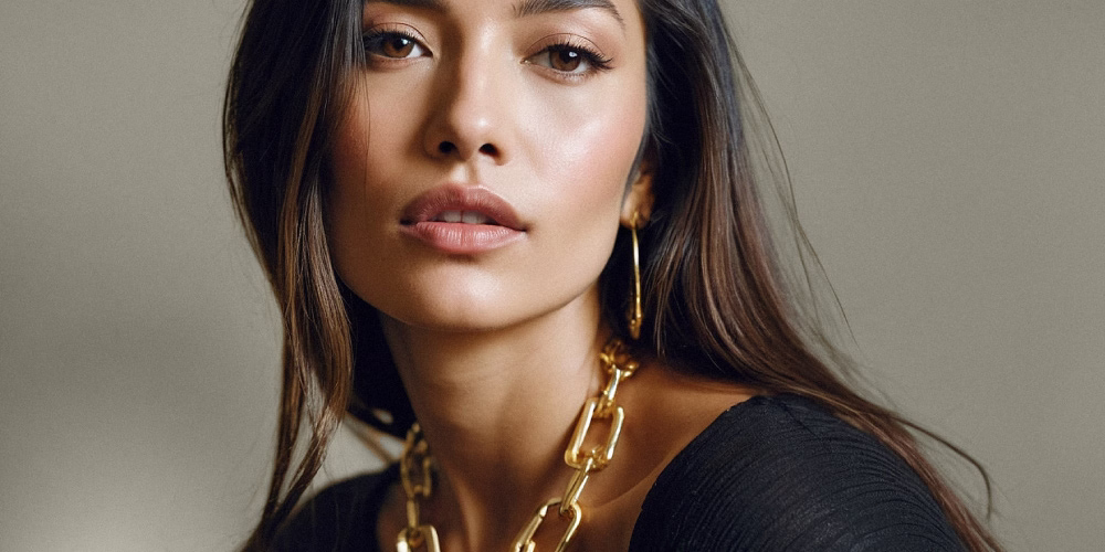A close-up portrait of a woman with long, dark hair, wearing a black top and large hoop earrings, alongside a chunky gold chain necklace.