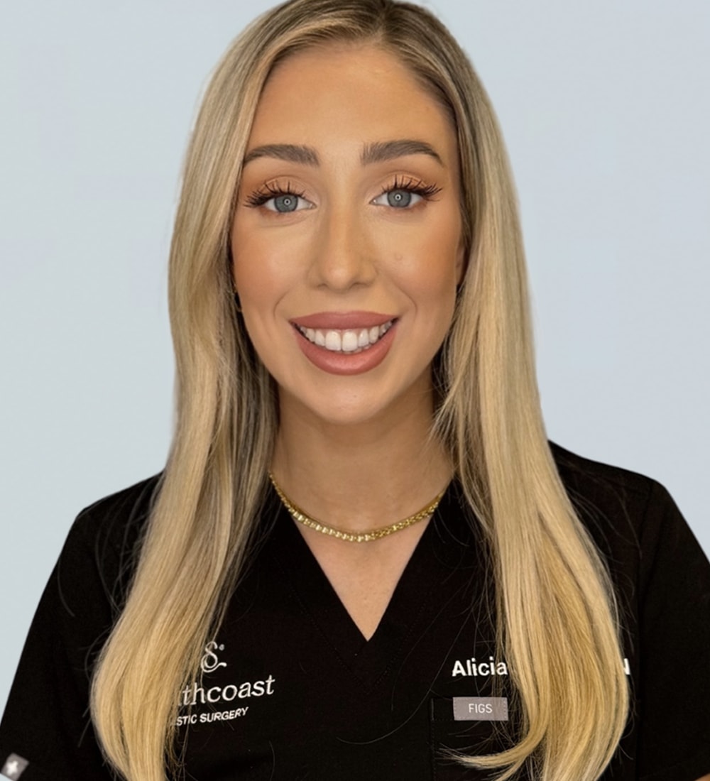 A woman with long blonde hair wearing a black medical uniform featuring the text 'Southcoast Plastic Surgery' and her name 'Alicia' on the left side, along with the word 'FIGS'.