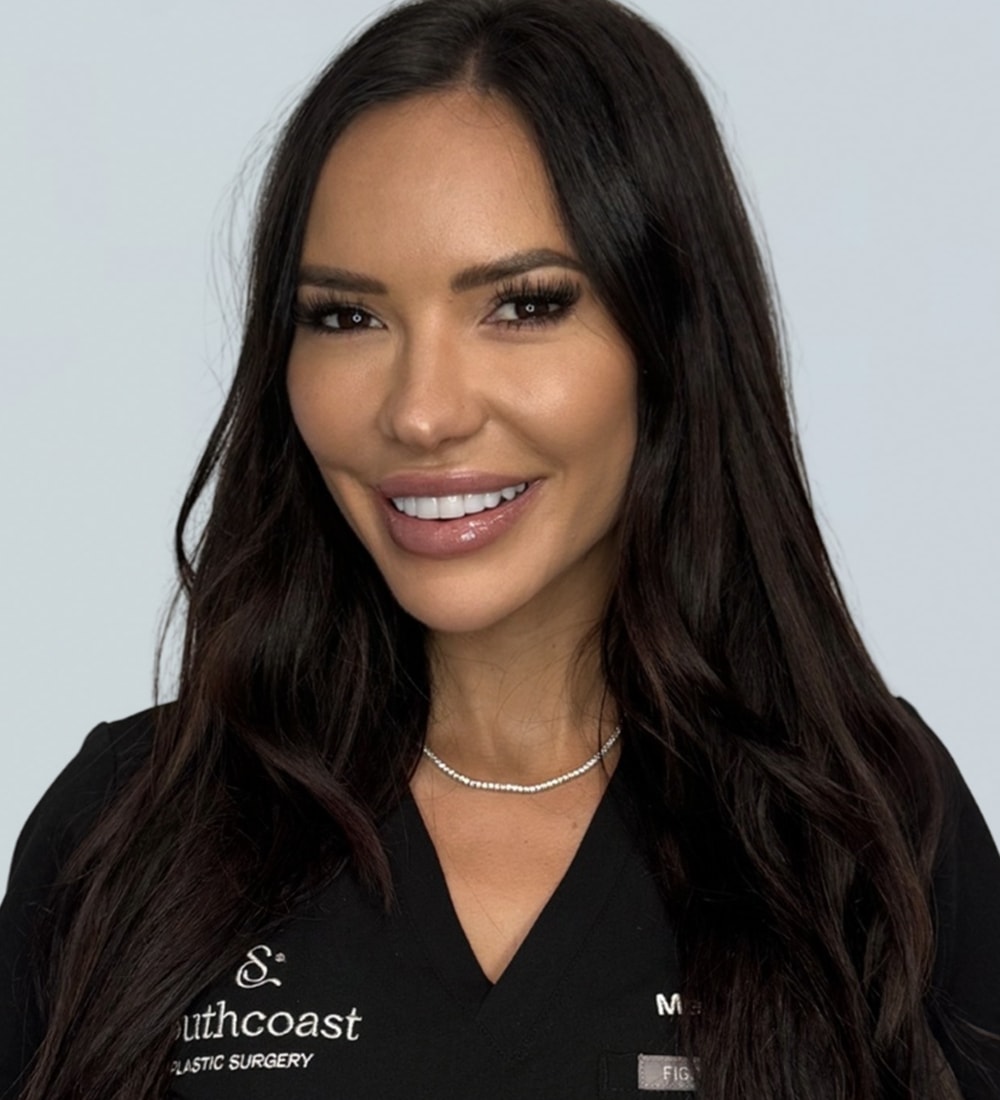 A woman with long dark hair smiling, wearing a black medical scrub top with the text 'Southcoast Plastic Surgery' visible on the left side.