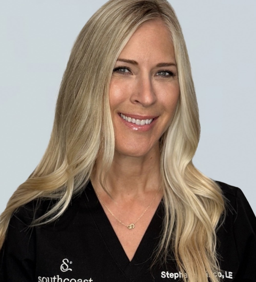A smiling woman with long blonde hair wearing a black medical scrub top, which has the text 'southcoast' and 'Stephanie, C.O.L.E.' printed in white.