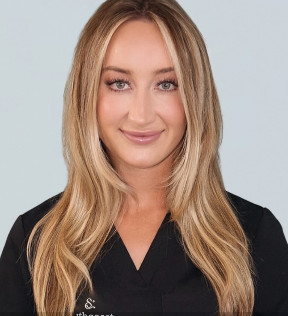 A woman with long blonde hair wearing a black medical scrub top, looking directly at the camera with a friendly smile.
