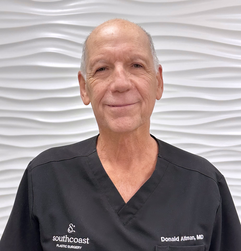 A man wearing a black scrub top with 'southcoast' and 'Donald Altman, MD' printed on it, smiling and looking directly at the camera.