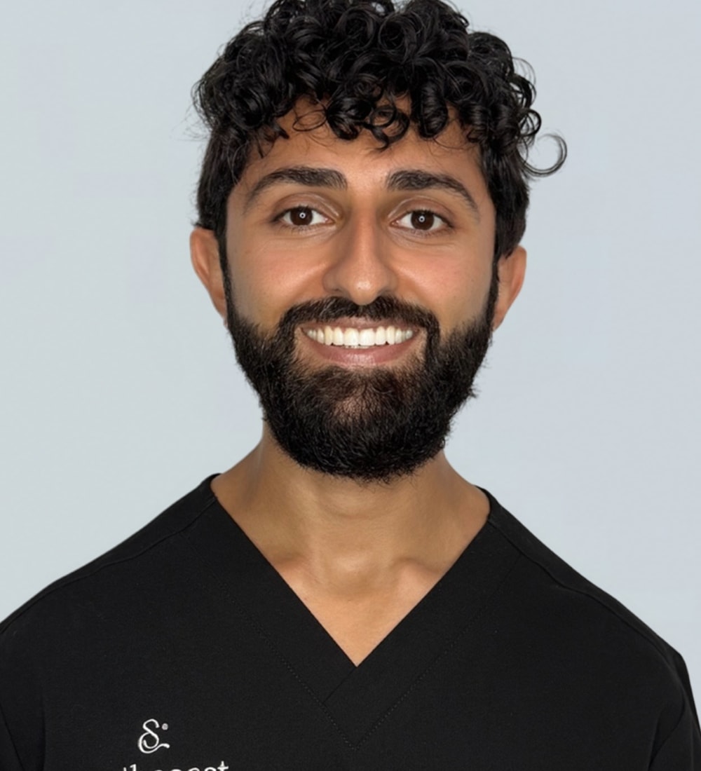 A smiling male with curly black hair and a beard, wearing a black scrub top with white text that reads 'S.' and 'the best.'