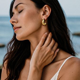 A woman with long dark hair, wearing gold hoop earrings, lightly touching her neck while looking peaceful against a blurred coastal background.