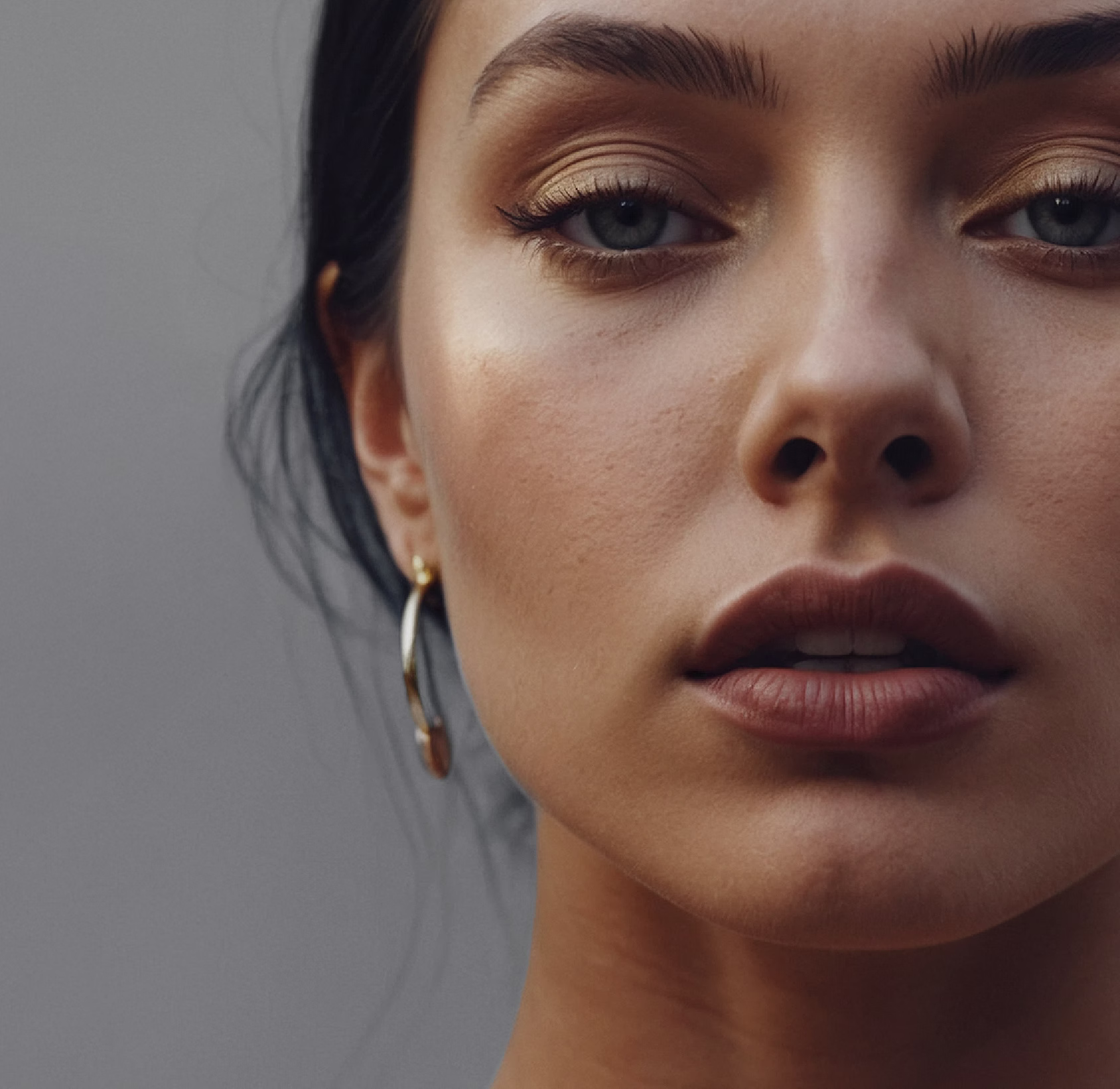 A close-up portrait of a woman with light makeup, featuring gold eyeshadow and natural lip color. She has long dark hair, wears a hoop earring, and has a neutral expression.