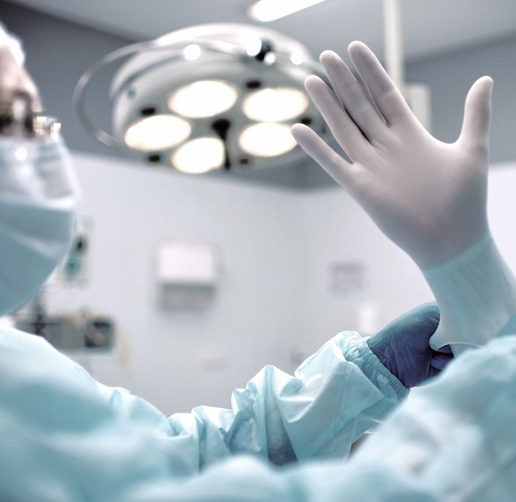 A person in a surgical mask and gloves is putting on a sterile glove with their right hand, while raising their left hand with the palm facing up.