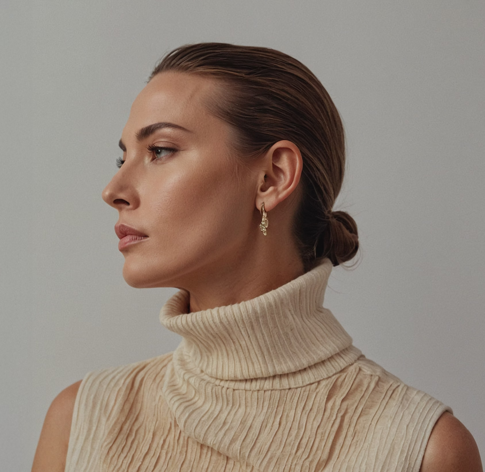 A profile view of a woman wearing a high-neck, ribbed cream sweater, showcasing elegant, dangling earrings while her hair is styled in a low bun.