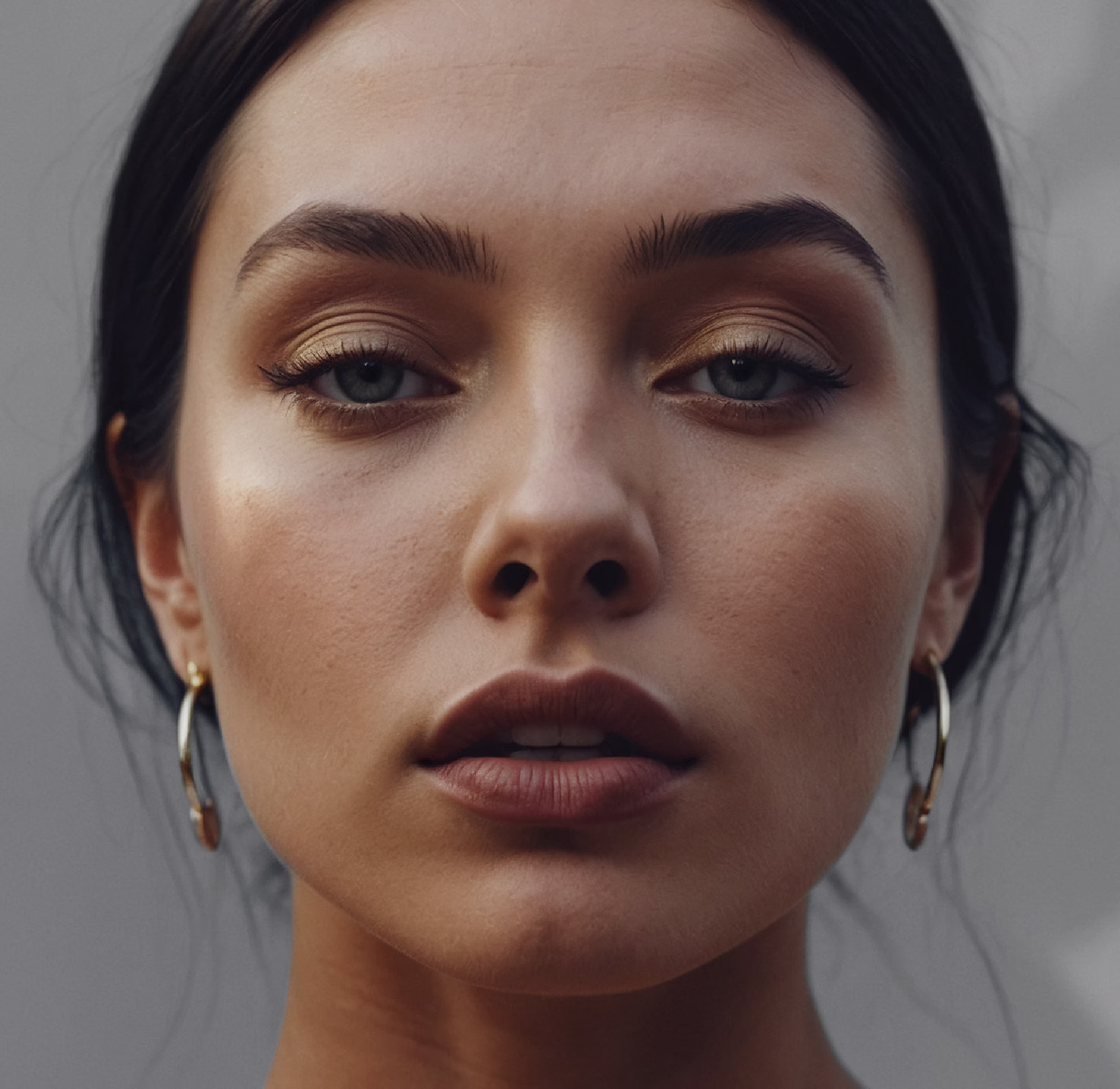 A close-up portrait of a woman with long dark hair, wearing gold hoop earrings. She has a neutral expression, with defined brows and natural makeup, highlighting her features.