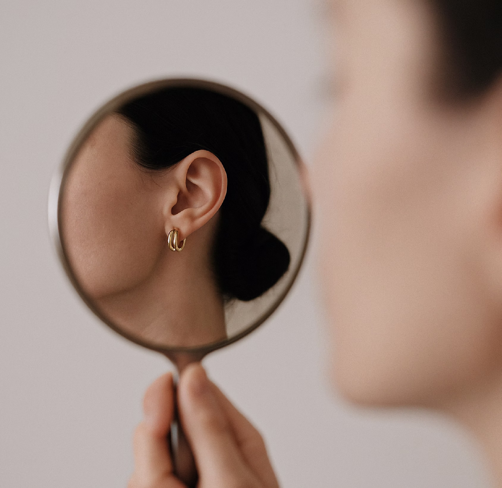 A woman holding a round mirror reflecting her profile, showcasing a gold hoop earring in her ear.