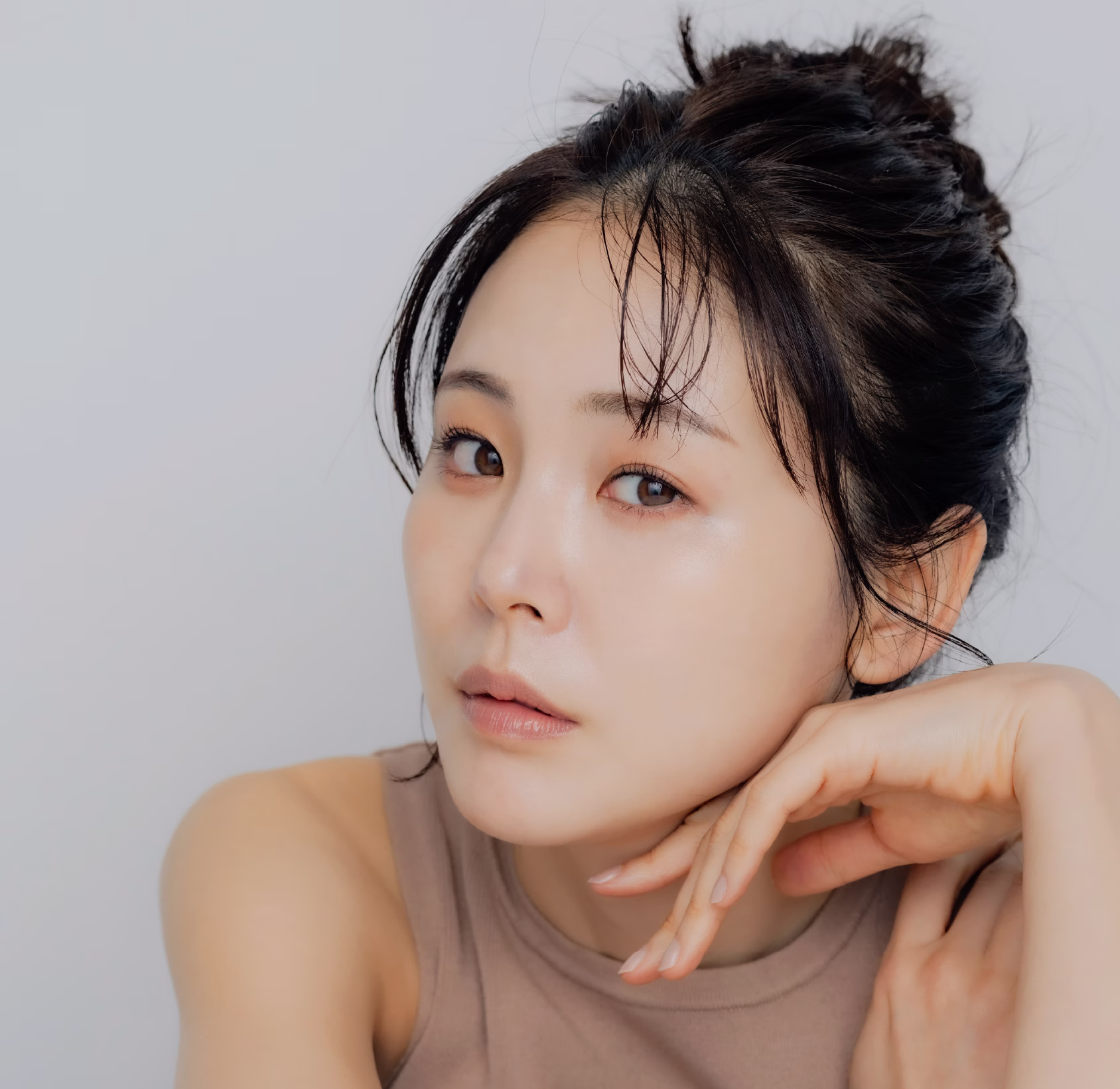 A woman with dark hair styled in a messy bun, wearing a sleeveless, light-colored top, resting her chin on her hand while looking directly at the camera with a natural expression.