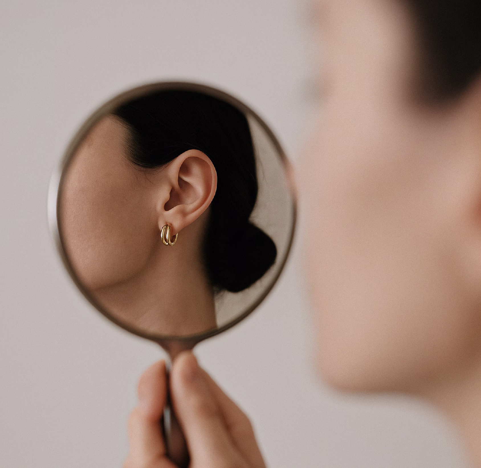A woman holding a small round mirror reflecting her ear with a gold hoop earring.