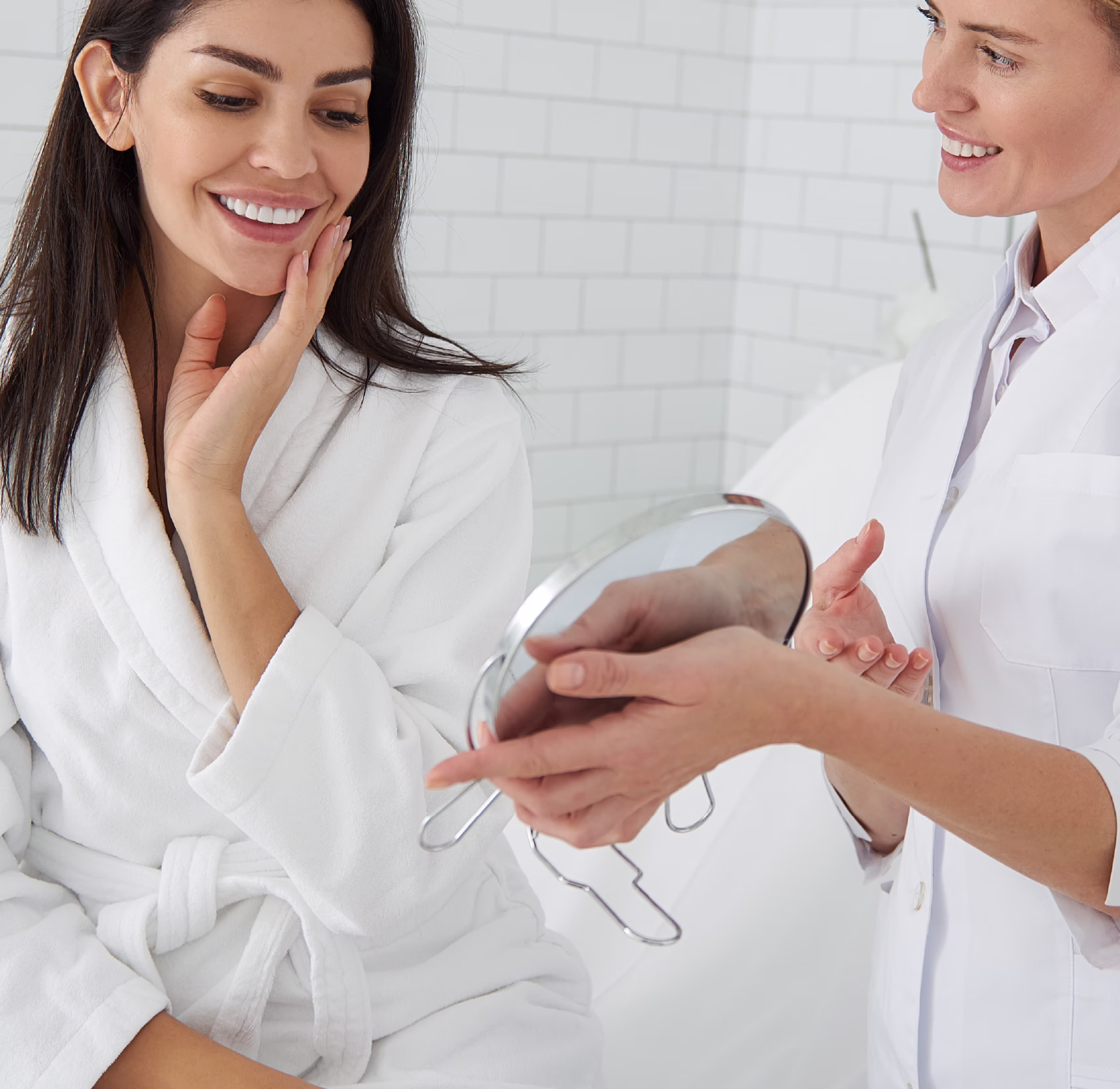 A woman in a white robe smiling while touching her face, and a woman in a white coat holding a round mirror, both engaged in conversation.