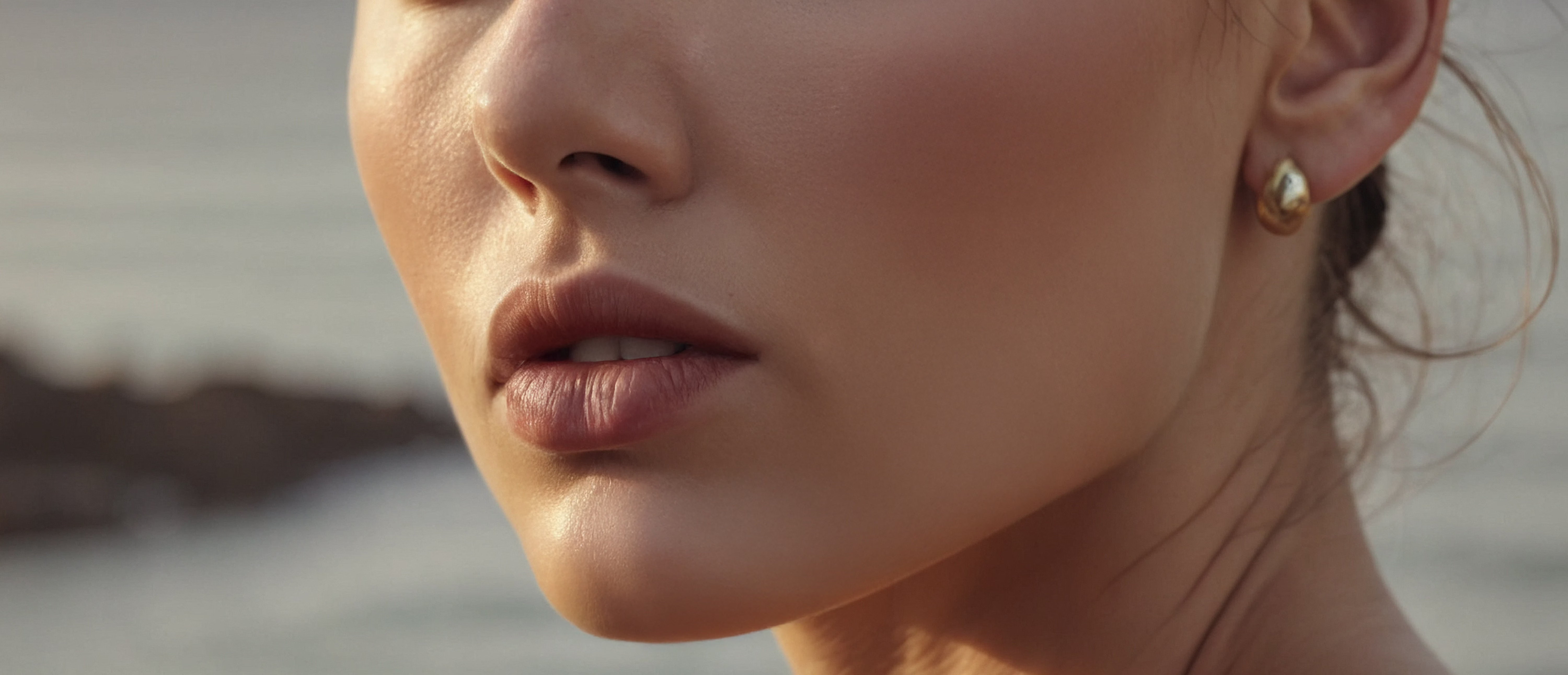 A close-up of a woman's face, focusing on her lips and jawline. She has a natural makeup look with soft skin tones and is wearing a small gold earring. The background is softly blurred, highlighting her facial features.