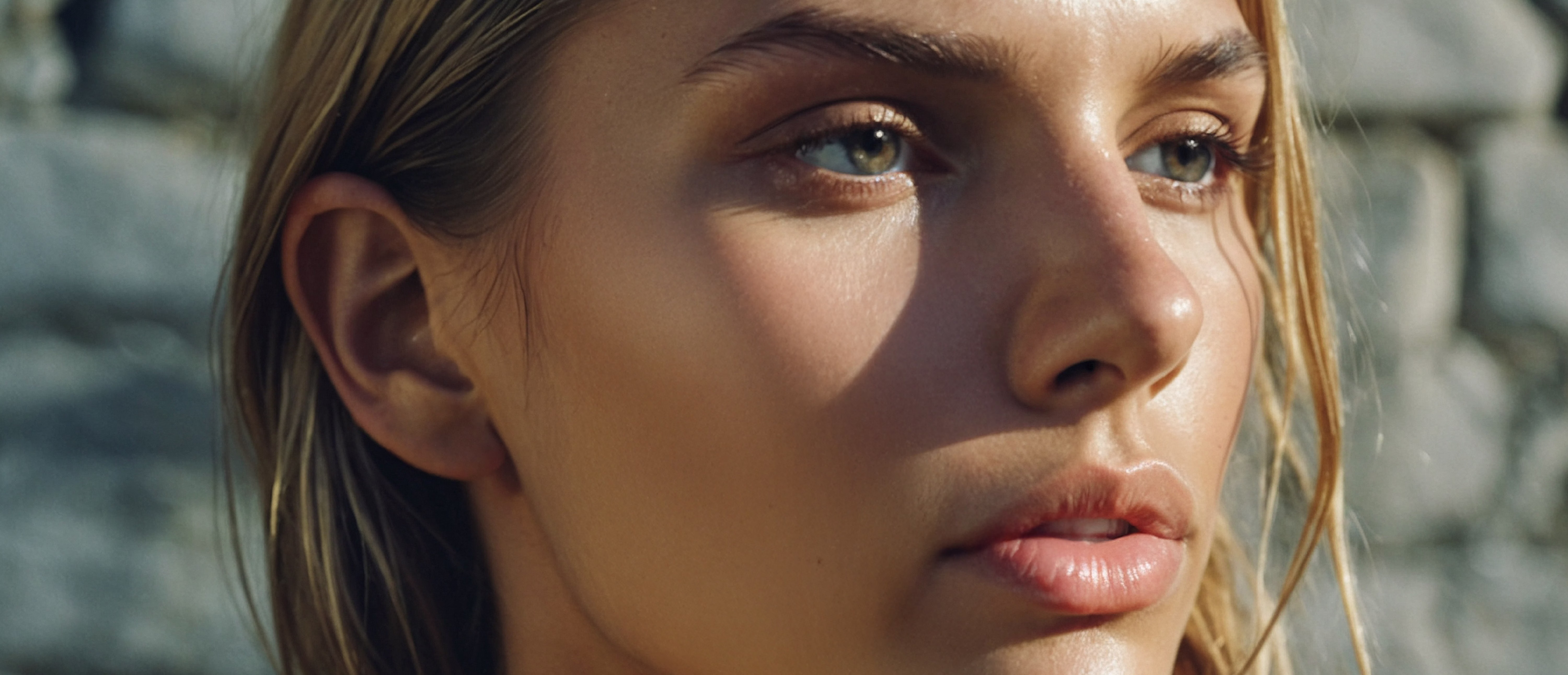 A close-up portrait of a young woman with light brown hair and blue eyes, featuring a natural makeup look. She has a soft expression, and sunlight highlights her facial features.