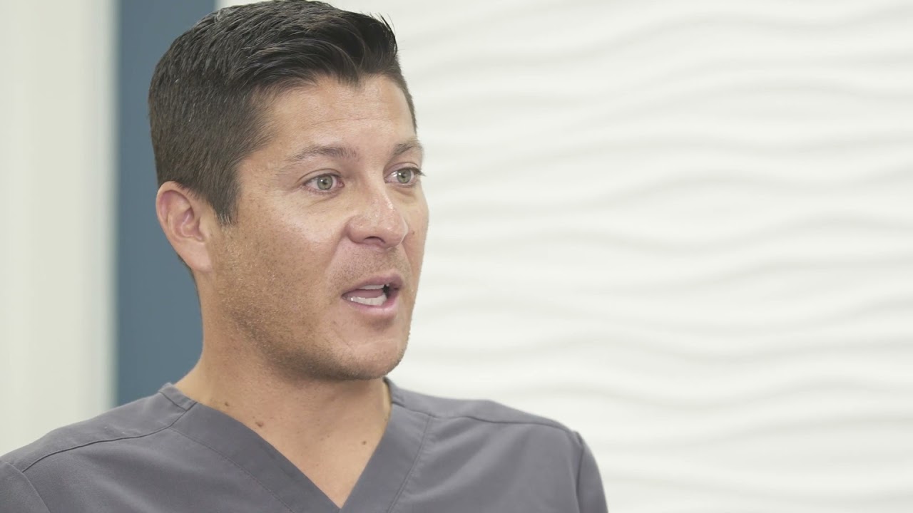 A man wearing a gray medical scrub top is speaking, with short black hair and a serious facial expression.