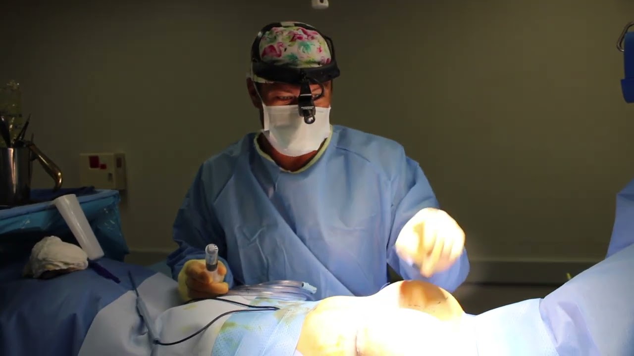 A surgeon wearing a floral-patterned surgical cap and a face mask, using a surgical instrument while standing beside a patient on an operating table, dressed in surgical scrubs.