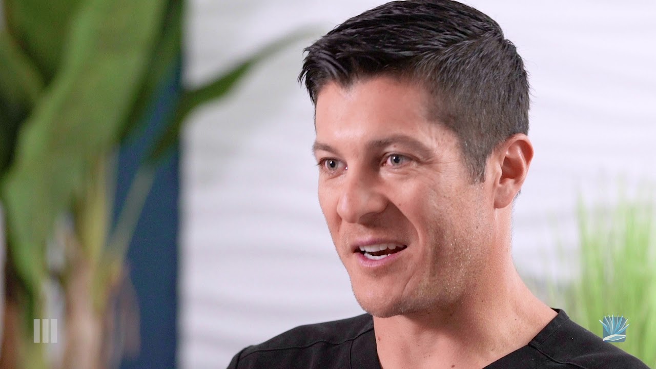 A person with short black hair and wearing a black shirt is smiling while talking. They have a friendly expression and are positioned to the right of the image.