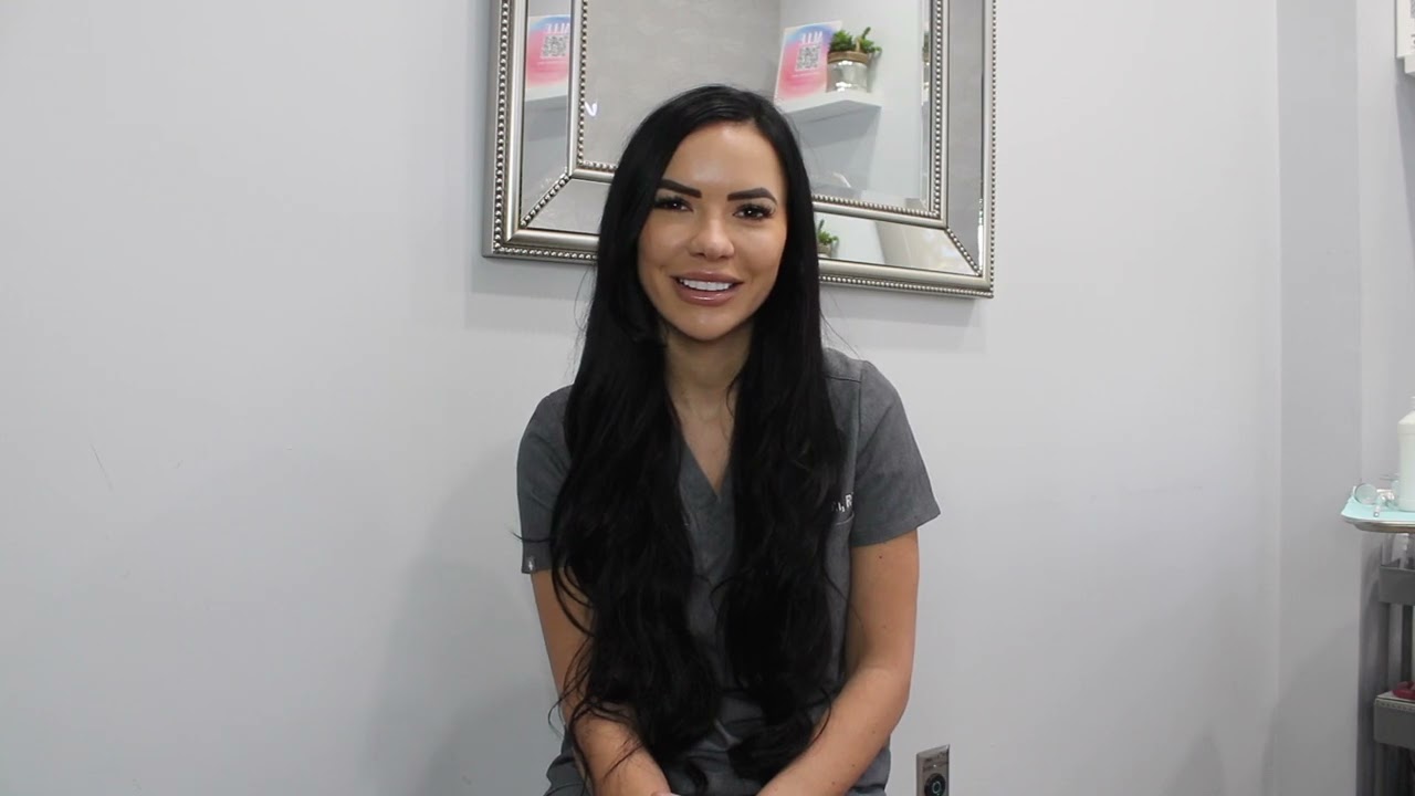A woman with long black hair, sitting and smiling, wearing a gray outfit. She is in front of a decorative mirror.