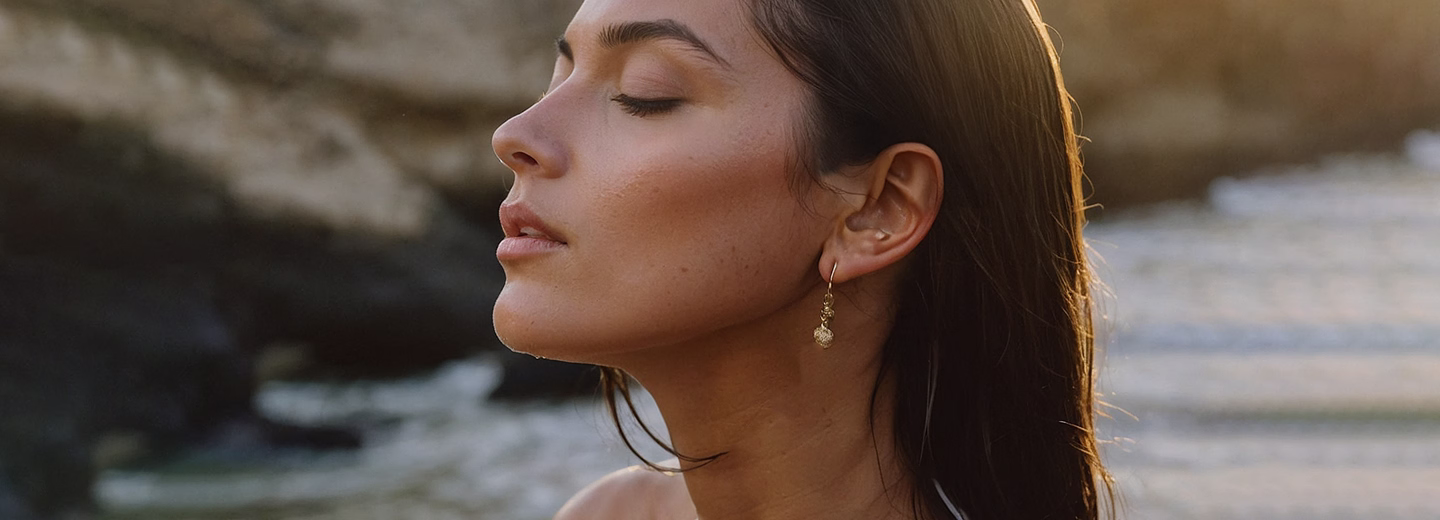 A woman with long hair is closing her eyes and breathing deeply, wearing a gold earring, with sunlight softly illuminating her face.