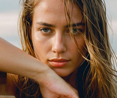 A close-up portrait of a woman with wet, wavy hair, gazing directly at the camera with a serious expression. Her hand rests on her chin, and she has a natural makeup look showcasing her features.