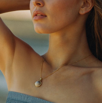 A close-up image of a woman wearing a delicate gold necklace featuring a round pendant with a blue and white iridescent surface.
