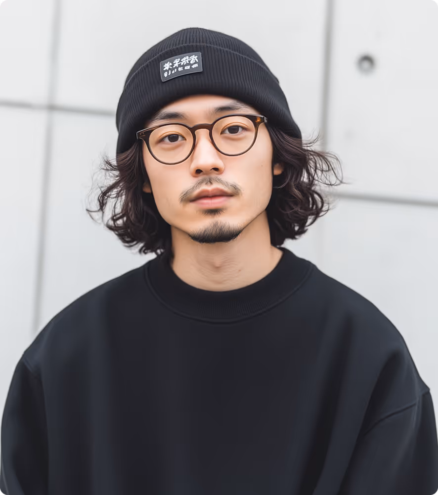 Young man with curly hair wearing round glasses, a black beanie, and a black sweatshirt standing against a tiled wall.