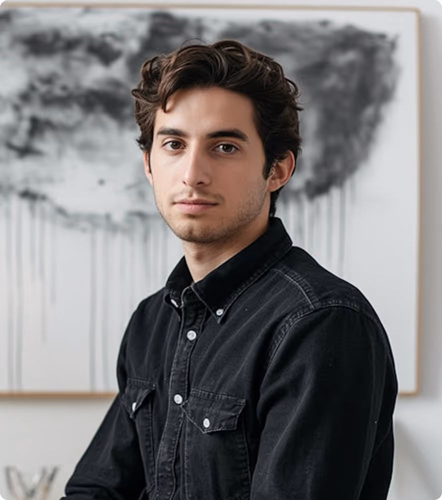 Young man with dark curly hair wearing a black button-up shirt, sitting in front of a blurred abstract black and white painting.
