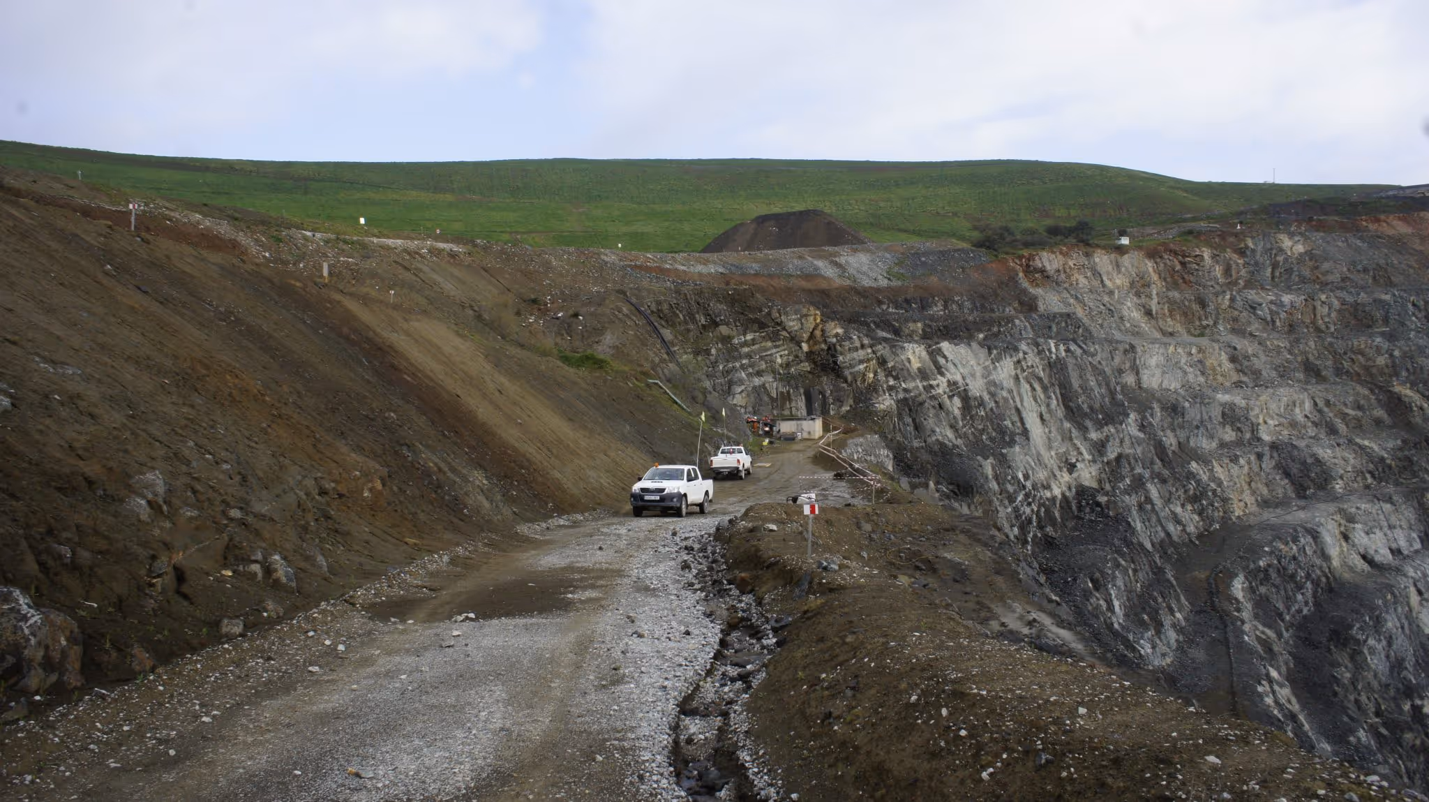 Camionetas blancas circulando por un camino de grava en una mina a cielo abierto con terreno rocoso y una colina verde al fondo.