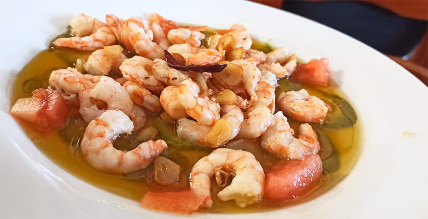Plato de gambas al ajillo con ajo dorado y trozos de tomate en aceite.