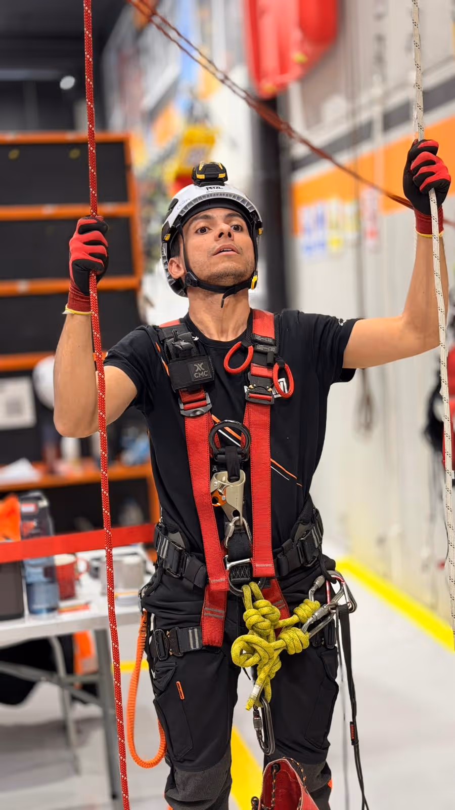 Hombre con casco y arnés de seguridad manipula cuerdas en ambiente industrial interior.