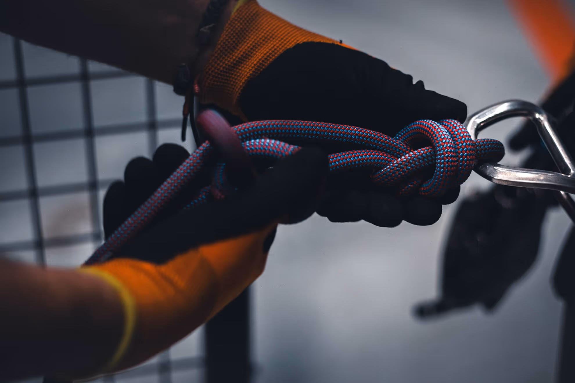 Manos con guantes negros y naranjas sujetando una cuerda azul y roja con nudo, enganchada a un mosquetón metálico.