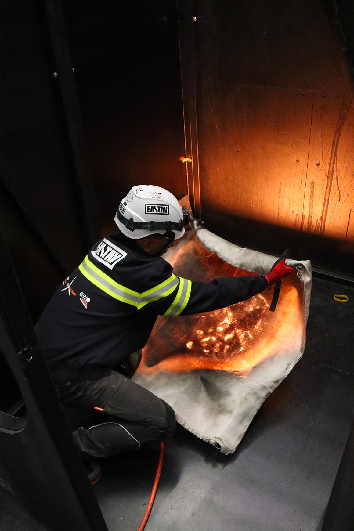 Trabajador con casco y ropa de protección sostiene una manta contra incendios parcialmente prendida fuego en una habitación oscura.