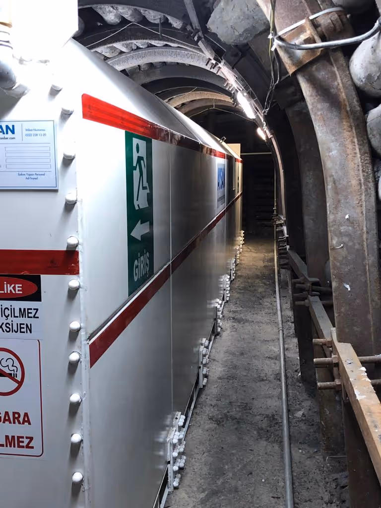 Pasillo estrecho industrial con señalización verde y roja, tuberías oxidadas y equipo pesado.