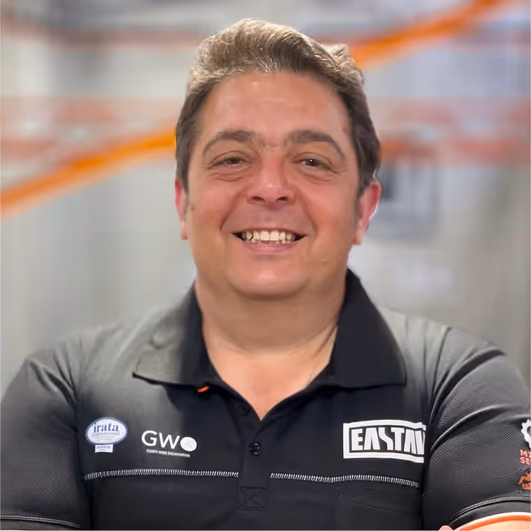 Hombre sonriendo vistiendo polo negro con logos GWO y EASTN en fondo borroso.