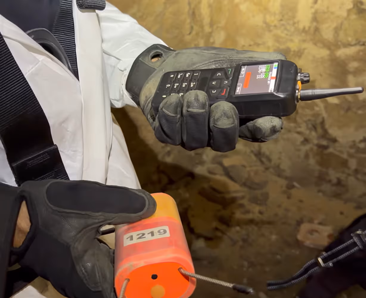 Persona con guantes sostiene un dispositivo negro con pantalla y botones junto a un cilindro naranja marcado con el número 1219.