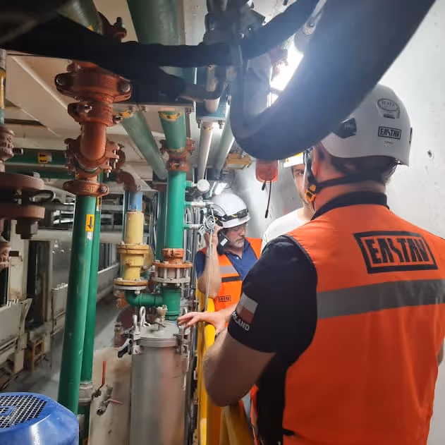 Tres trabajadores con cascos y chalecos naranjas revisan tuberías y maquinaria en una sala de equipos industriales.