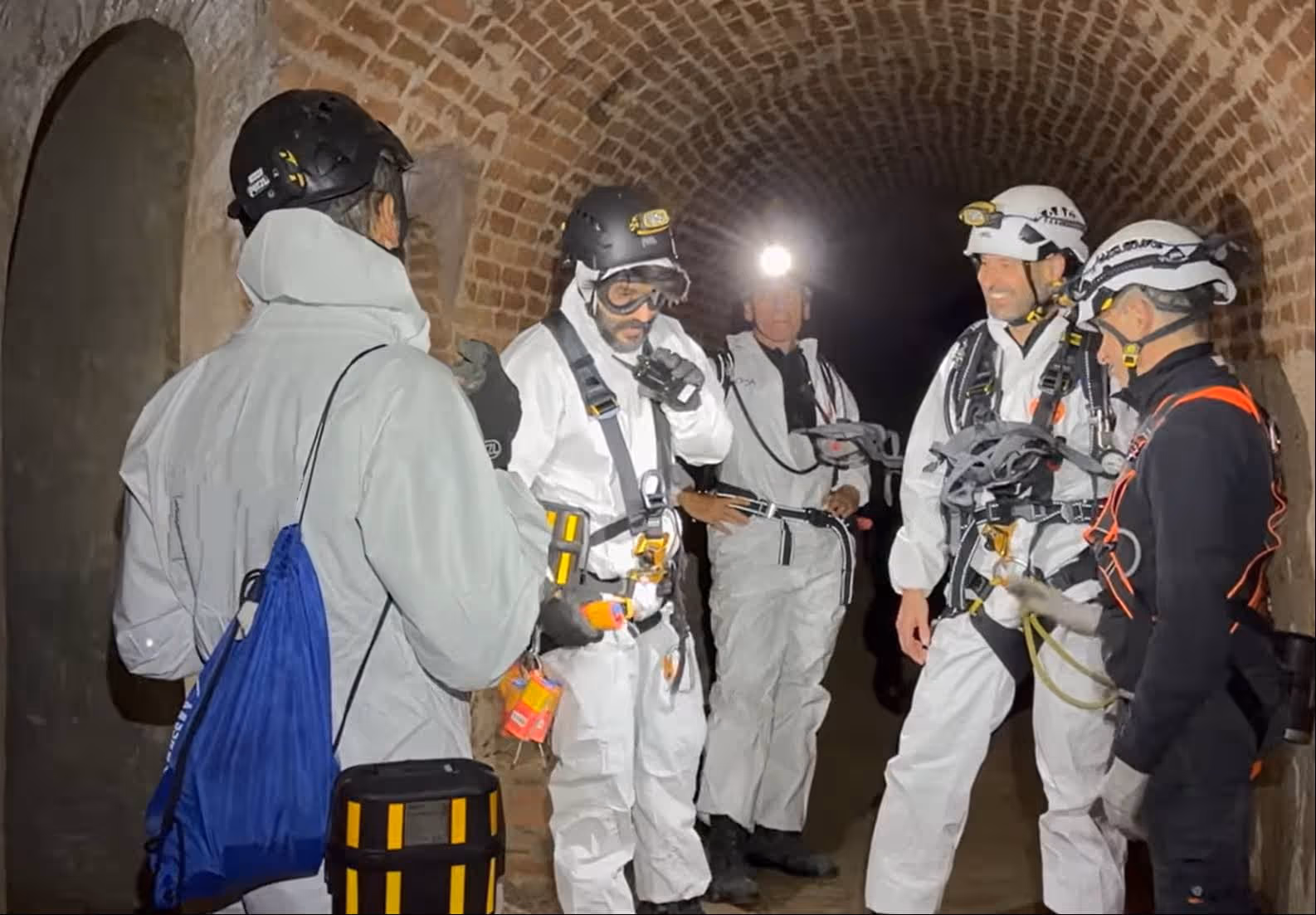 Un grupo de cinco personas en un túnel de ladrillo con equipo de protección, cascos y luces frontales, preparándose para una exploración o inspección.