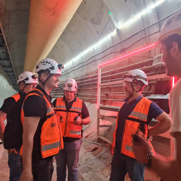Trabajadores dentro de un túnel con chalecos reflectantes naranjas y cascos blancos, rodeados de iluminación roja y blanca.