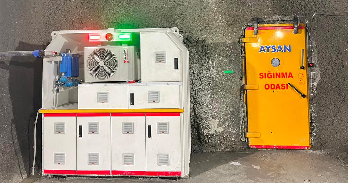 Puerta amarilla de refugio de emergencia en pared de roca con letrero rojo y azul que dice 'Sığınma Odası' y equipo técnico blanco al lado con luces roja y verde.