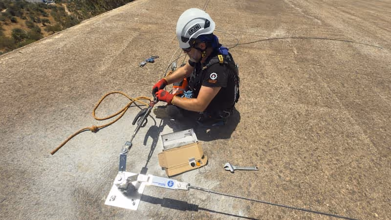 Trabajador de seguridad equipado con casco blanco y guantes rojos instalando un anclaje de seguridad en una superficie de concreto.