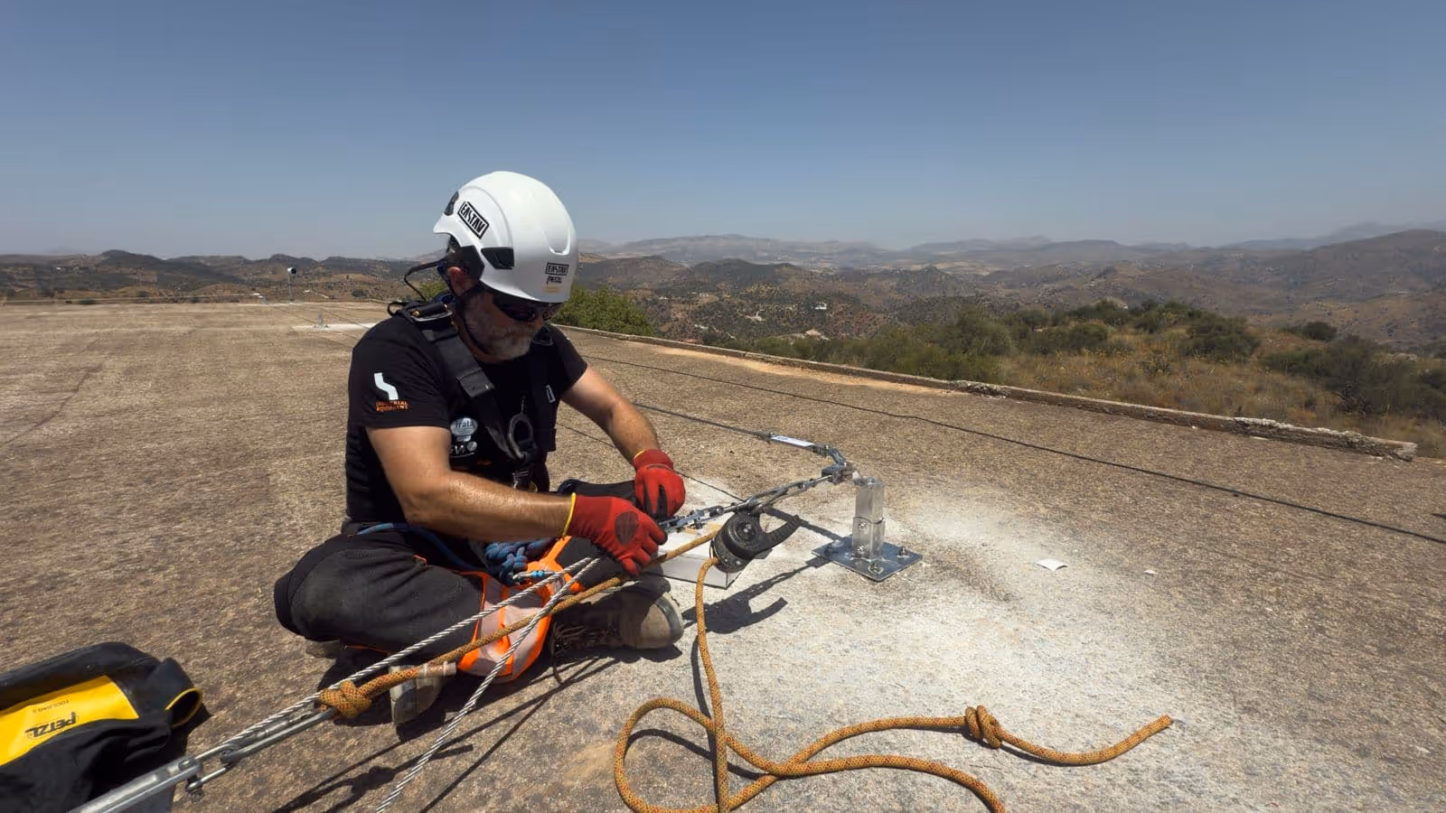 Trabajador con casco blanco y guantes rojos asegura cuerdas en una superficie de concreto al aire libre con montañas al fondo.
