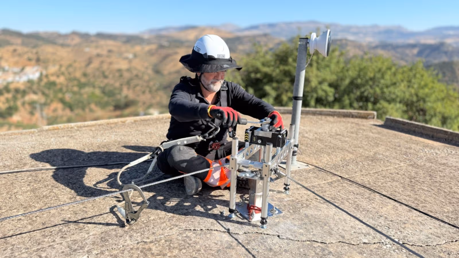 Trabajador con casco y guantes ajustando equipo técnico de metal asegurado con cables en una azotea al aire libre.