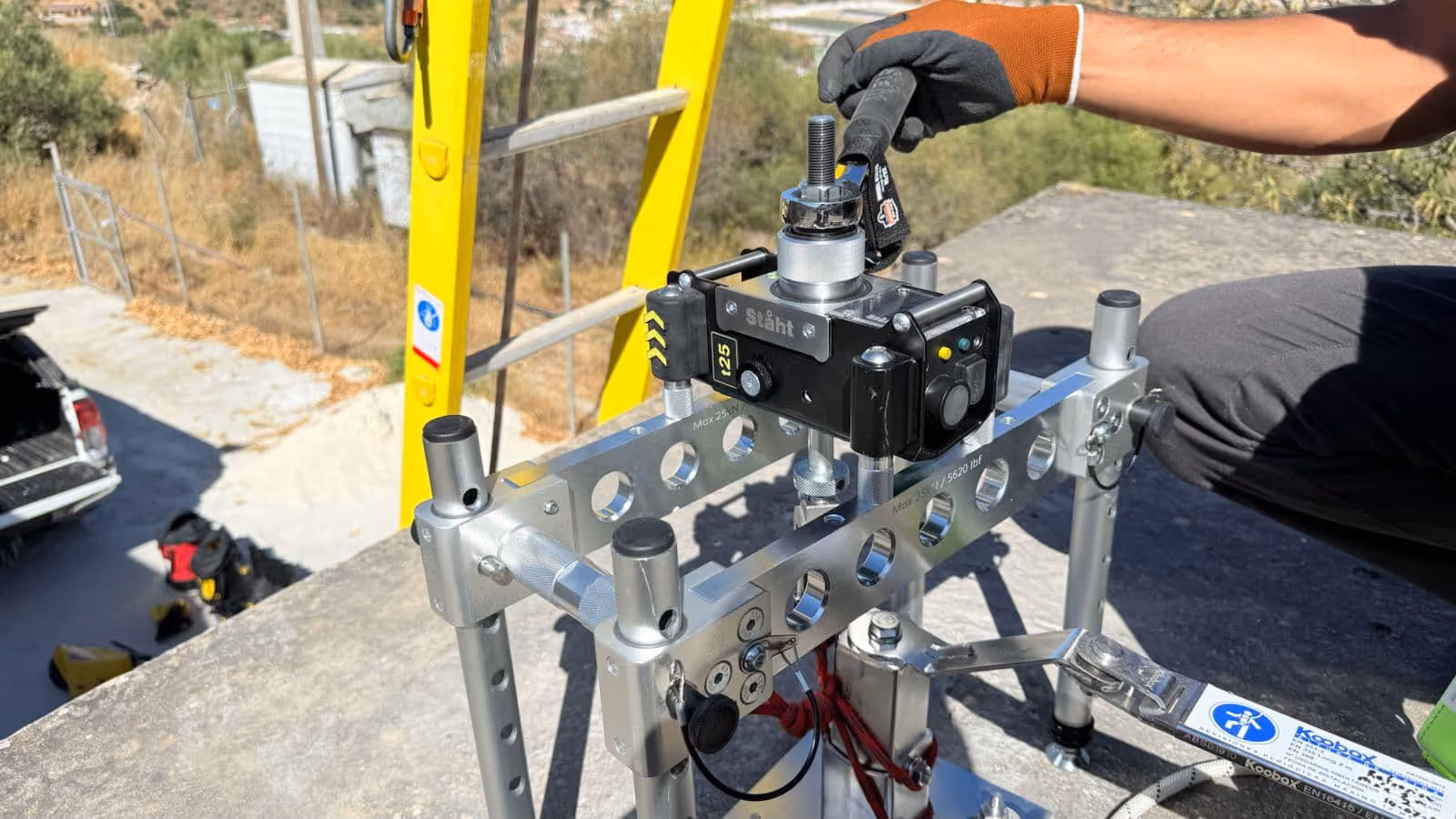 Mano con guante naranja ajustando un equipo de medición de torque negro montado en una estructura metálica con fondo de exteriores y escalera amarilla.
