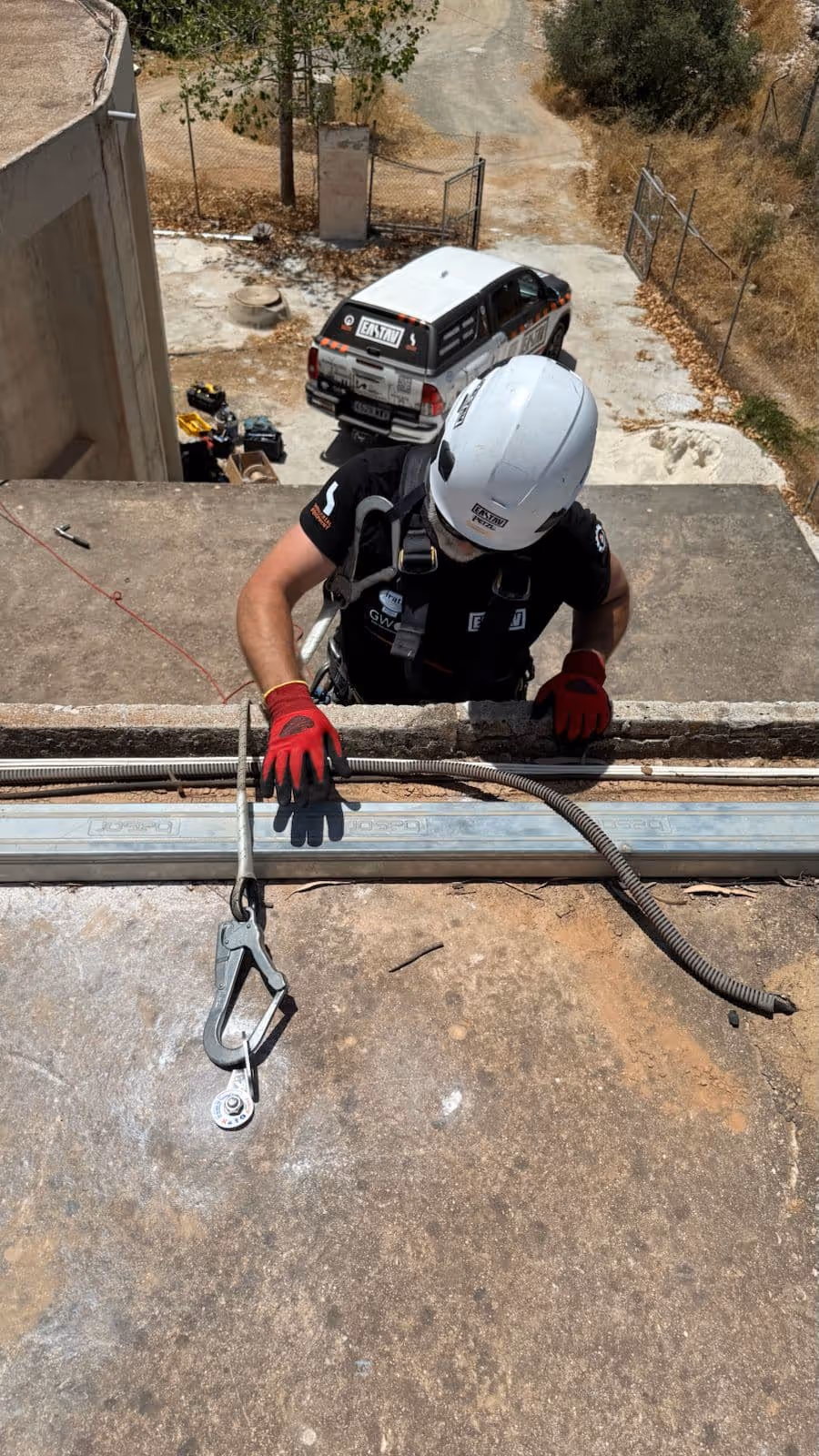 Trabajador con casco blanco y guantes rojos asegurado con arnés mientras escala un muro de concreto en un entorno industrial.