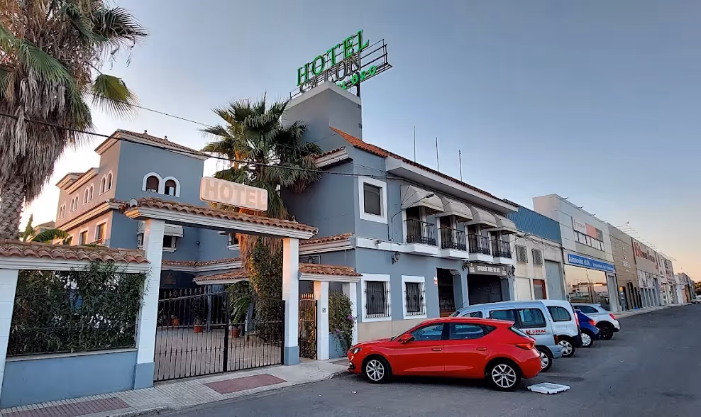 Edificio azul de hotel con letrero verde luminoso en la azotea y varios coches estacionados frente a la entrada.