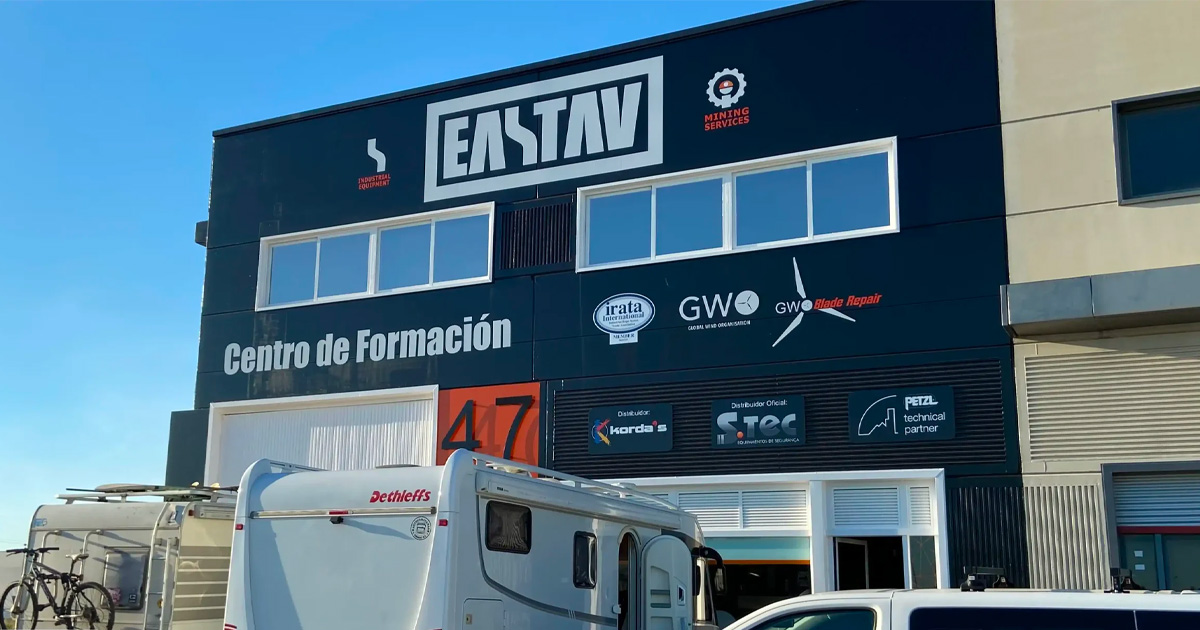 Fachada de un edificio industrial negro con el letrero 'EASTAV Centro de Formación' y varios logos de empresas asociadas, con vehículos estacionados enfrente bajo un cielo azul.