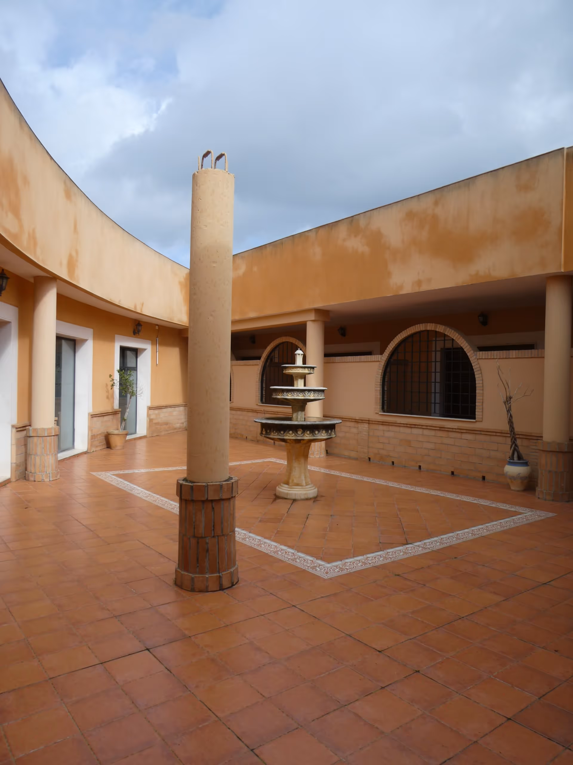 Patio interior con suelo de tejas naranjas, columna central, fuente de tres niveles y paredes color terracota con ventanas arqueadas.