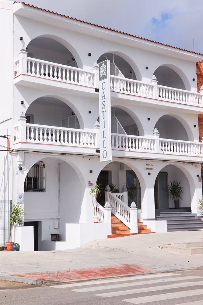 Fachada blanca de tres pisos con arcos y barandales, con un cartel vertical que dice 'Castillo' en un hostal.