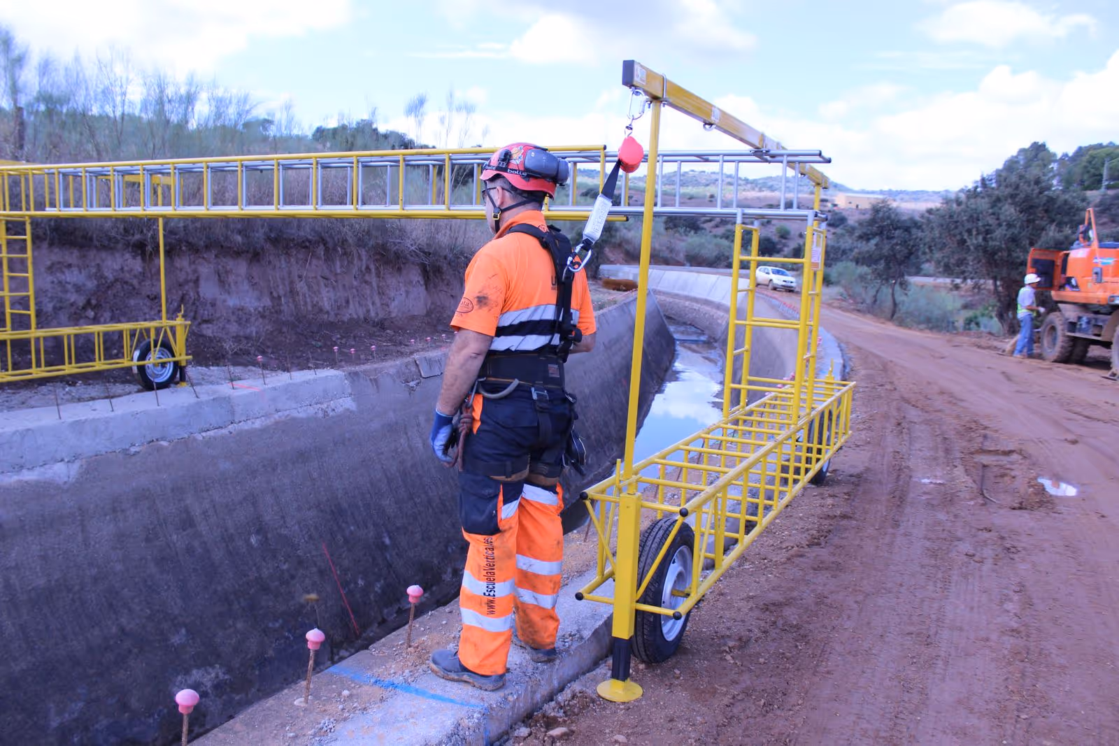 Trabajador de construcción con arnés de seguridad conectado a una estructura amarilla móvil al borde de un canal de concreto en una zona de obra.