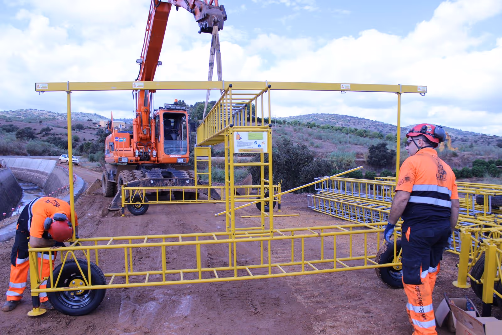 Dos trabajadores con ropa de seguridad instalando una estructura amarilla móvil en un terreno con maquinaria pesada naranja de fondo.